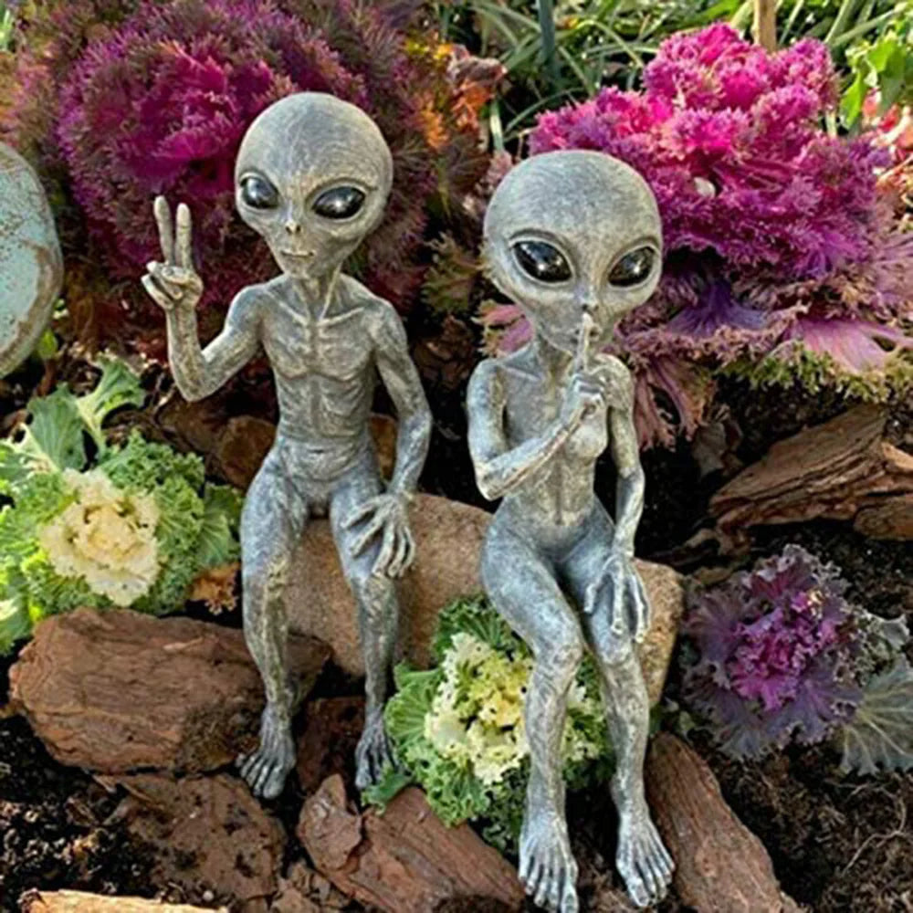 Two alien figurines in a garden setting with flowers and rocks.